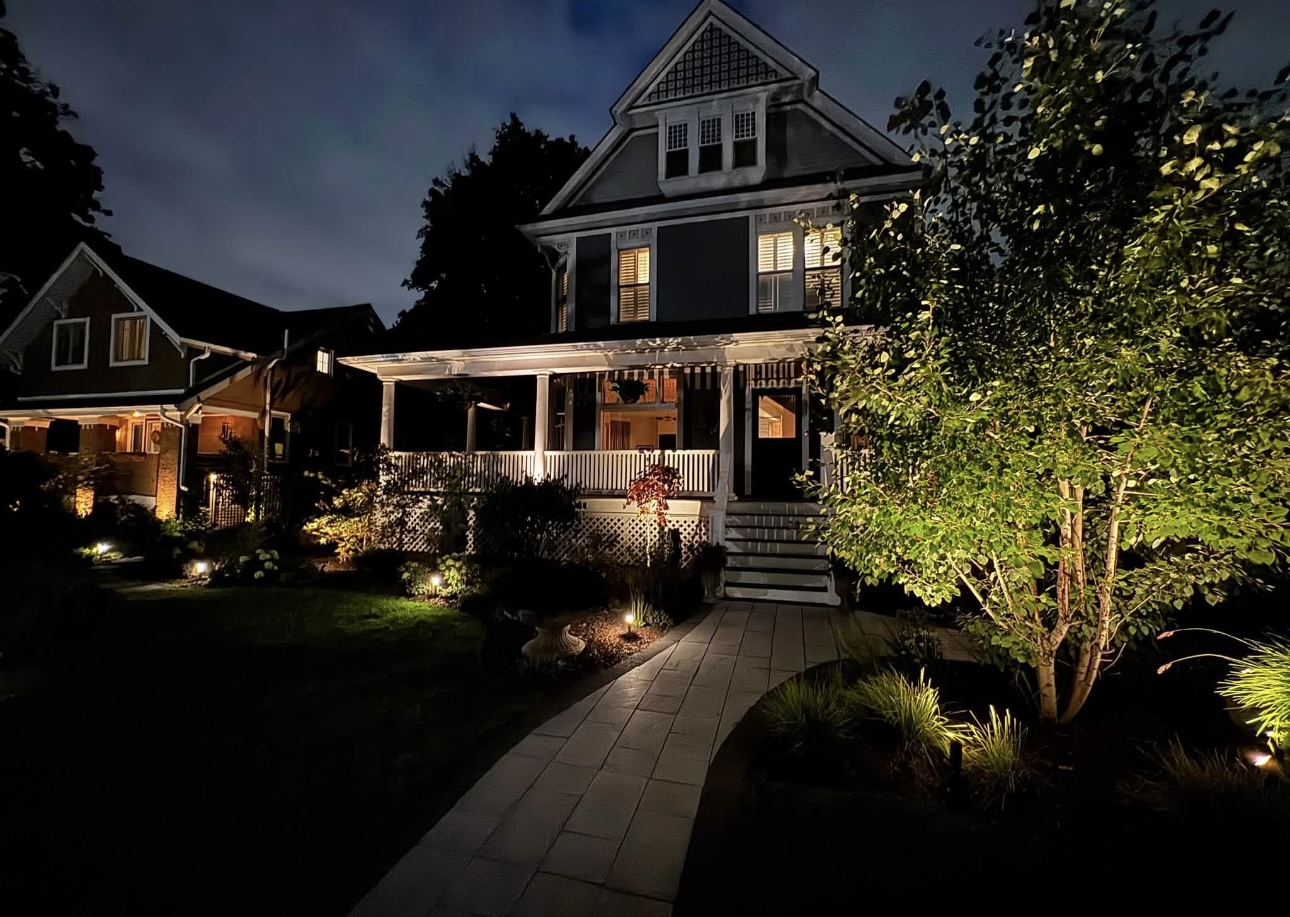 Dark gray home with warm white porch, walkway, and tree lighting under a cloudy night sky