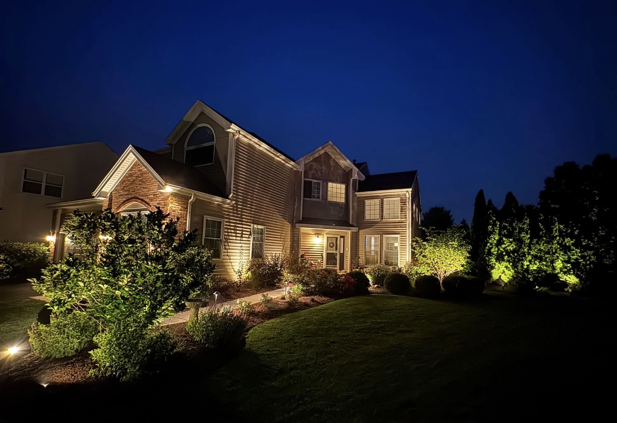 Beige and brick home with warm white lights under the eaves and uplighting the walkway and trees