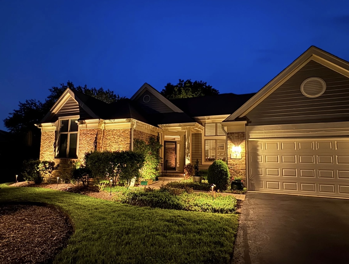 Tan brick and gray siding home with warm white landscape and walkway lighting at twilight