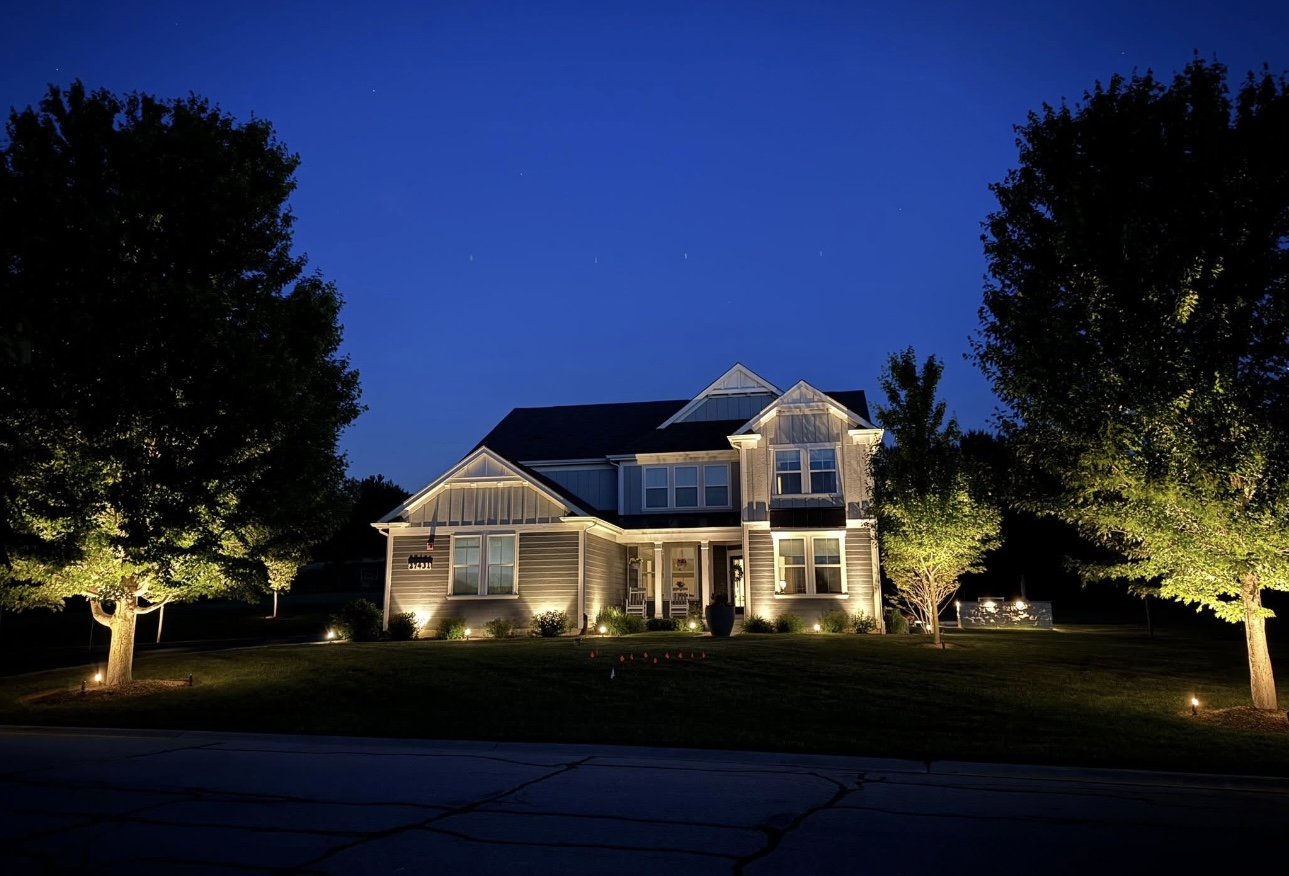 Gray home with warm white light washing the lower walls and yard trees at dusk